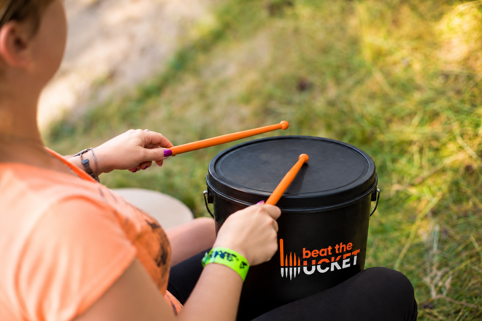 A participant using drumsticks to play a black Beat The Bucket pail outdoors.